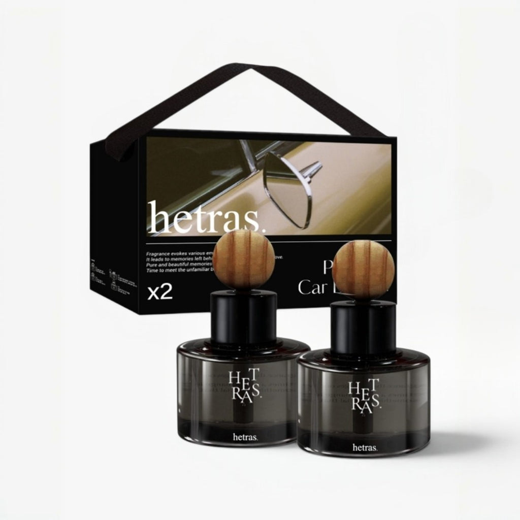 Two Het Ras car fragrance bottles with round wooden tops displayed in front of their black presentation box on a light gray background.